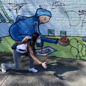 Photo of a person with long dark hair and a white mask on standing in a crouched position imitating the mural behind them of a person reaching for a vessel