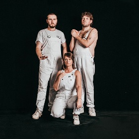 Photo of three artists in white bib and brace overalls, two standing one crouching, all make hand gestures