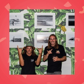 Photograph of two people with long hair and moustaches, making rock on symbols with hands. One wears a bucket type green hat. Behind them is a series of black and white  surfing photos  and behind that is a green wallpaper of banana leaves / palms