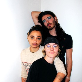 Photo of three people embracing and posing for the photo, one wears round glasses and a beanie they have curly short blond hair, one wears a long sleeve white t-shirt and looks like they have braided hair pulled back, one at the back has long dark hair facial hair and blue eyeshadow