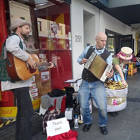 Photo of 3 street performers, one has a guitar and wears a little hat with a brim, one has a paino accordian and the other is bent over and it is hard to see what they are doing, the image is of them standing in front of a shop mid performance
