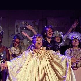 Promotional image for Creation, elaborately dressed performers are on stage, singing, smiling and dancing, there are many gold tones and glitter