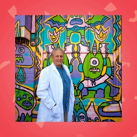 Photo of a person in a white lab like coat and blue shirt, they stand in front of a  multicoloured graffiti wall