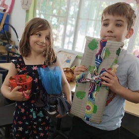 Two children pose for a photo, one on the left with shoulder length hair wearing a dress smiles cheekily glancing at the other who is in the forefront they have short hair and wear a grey tshirt, both hold hand made gifts wrapped in recycled materials