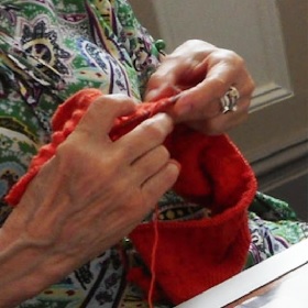 Photo of two hands knitting with orange wool, there are silver rings on the left hand and the person wears a paisley green and blue  top