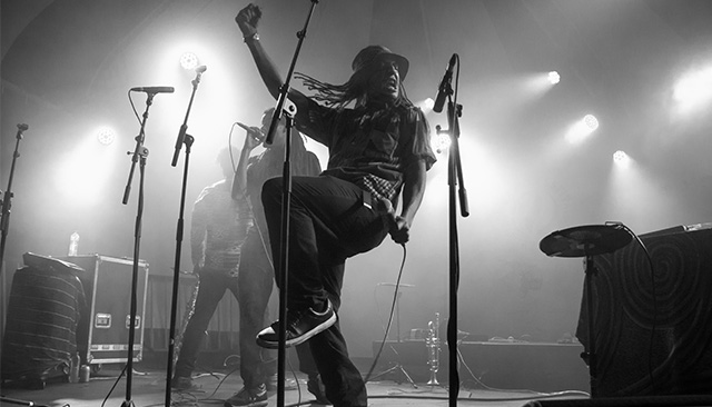 Black and whote photo of Vuli on stage, he has one arm raised above his head, and one leg bent at the knee and raised, his other hand holds a mic, its an action shot and his brother can be seen behind him singing into the mic, the third band member can also be seen on stage. Bright lights shine down on the band while they perform