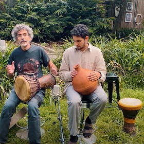 Two musicians sit in a garden, one has a drum on their lap the other has a clay pot they are hitting on their lap.