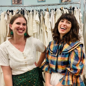 Photo of two stylish people posing and grinning at the camera, behind them is a rack of white clothes 