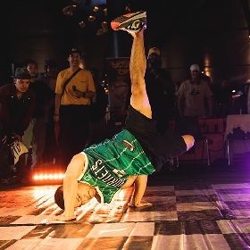 Photo of a street dancer in a semi handstand on a chequered black and white floor one leg points uo the the sky and the other is bent behind them. The dancer wears a green basketball singlet and black short, they wear sneakers, there are lights shining and it looks like night-time, an audience can be seen in the background