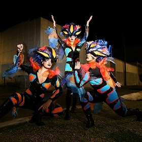 three street performers pose on the street, bright jester like bodysuits with large headdresses