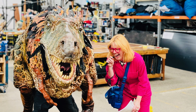 Photo of ERTH studio, a large yellow dinosaur puppet stands next to Amy Mills from Accessible Arts who wears an electric pink jumpsuit and laughs