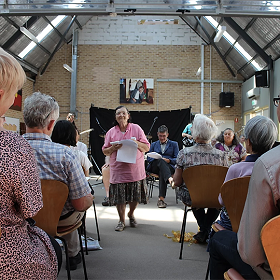Bold Stories promotional image, photo of woman in  floral skirt and pink shirt standing in front of audience  holding sheets of A4 paper in her hands