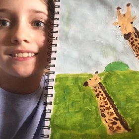 Photo of head and shoulder shot of child in pale blue t-shirt holding up an art book with a painting of two  giraffes in a field of green, grey sky in background of painting
