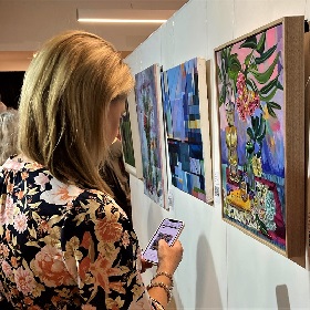 Photo of a person at a previous art show they look at their phone that has an image of the painting they are looking at on the wall