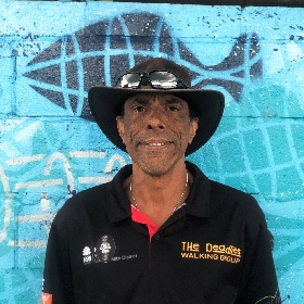 Photo of  Matthew Doyle, descendant of the Muruwari People, Matthew smiles and is wearing an Akubra hat and stands infront of a a aqua colour mural with a black fish stencil. His shirt reads the Deadlies walking group