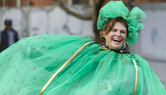 Photo of a person dressed in a green wearable art piece made from tafita, ther person has a very happy laughing oexpression on their face, the background is blurred and has a figure and a brick wall visible