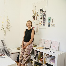 Photo of artist Nastia Gladushchenko inside the whites creek cottage studio, she leans against a low white bookshelf, the room has natural objects pasted on walls , neutral colours and wire, Nastia smiles warmly she is wearing a black short sleeved top and patterned pants also in neutral pinks and browns