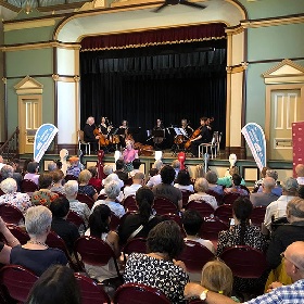 Photo of a classical band on stage at one of Council's town halls, there is a large audience in the foreground