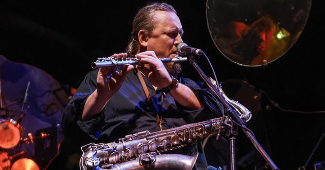 Musician James Ryan sits on stage with a saxophone on lap and pressing lips to a flute, he wears his hair in a ponytail and wears a black shirt and pants, the stage loos dark with lights on the performer