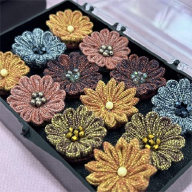 Photo of a box with what looks like knitted cord that has been designed into the shape of a flower, there are three rows of four flowers, made up of yellow, pink, orange and blue flowers with various beads in the centre of the flower