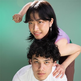 Photo of two young people, one wears a white t-shirt and looks at the camera, the other is leaning ontop of the first person with a menacing grin and raised fist with a large ring on it, the look off to the side