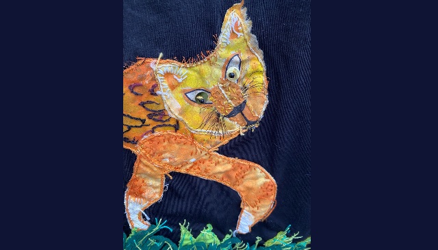 Tapestry of a kitten the cat is orange and has overlayed embroidery highlights and details in white, purple, orange, black and yellow. It also has sequin eyes and is standing on green grass tufts also material and embroidery. The visible part of the cat is its head, face and front two legs