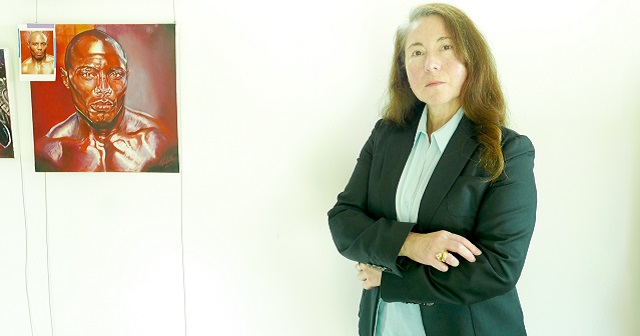 Photo of artist Miriam Cabello with arms crossed, wearing navy blue blazer and pale blue shirt, artist has long hair and stands in front of an artwork she has done of a person with brown skin and golden orangey tones, artwork is a square painting