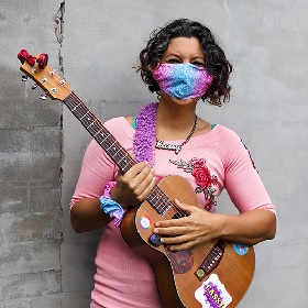 Photo of person against a wall wearing a light pink and blue mask with smiling eyes. Person has dark ear length wavy hair and a pink top. Person is holding a guitar with stickers on it