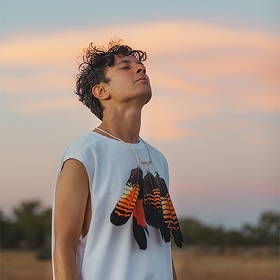 Photo of performer Dobby, he stands in an outdoor space they sky looks like a sunrise with light blues and soft pinks, he wears a white loose singlet and a feater necklace around his neck, his eyes are close and his face is held up to the sky