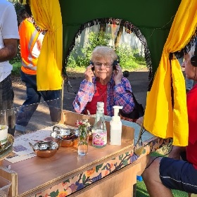 Photo of a person whit white hair sitting at the My Plate Your Plate wooden cart, they are polacing healphones on their head, in the foreground the wooden cart is visible with a rosewater bottlec, flowers and spices in metal bowls. 