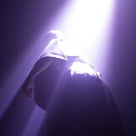 Promotional image for Ecstasy production at LIverworks, a body faces away from the camer with arms at their sides, they look like they are wearing a cape, there is a bright light and the image has no colour apart from a purple filter and the bright white lighting