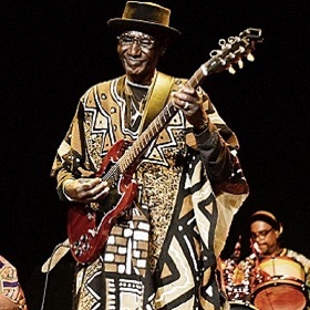 Performer Moussa Diakite on stage in hat and African traditional clothing, big smile and electric guitar in arms
