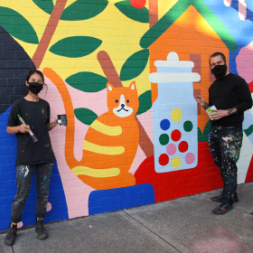 Photo of artist Kim Siew and Nico Nicoson in front of their mural All Sorts 2021, mural has a large ginger cat with yellow stripes and a white snout, there looks like a giant lollie jar containing round colourful sweets, there is also a house and a tree/foliage in the mural. The mural is in a simple, minimalist style with bold primary colours