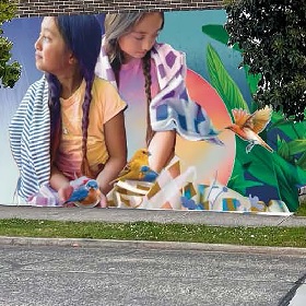 Photo of mural with two young girls with long plaits, both have scarves draped on their shoulder and both hold small blue and yellow birds on their palms there are leafy plant fronds to the right of the image and tha background is a white graduated colour to blue 