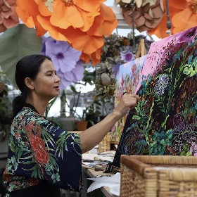 Photo of a person painting at an easel, their painting is very colourful and floral, they wear long hair tied back in a ponytail and a kimono-like dark blue, red, white and green top
