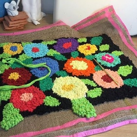 Photo of a black looking rug that has been embellished with lots of bountiful full flowers, green, red, blues, yellows, purples, oranges, pinks and reds. The rug sits ontop of a textured like matt which has earthy colours in a line pattern