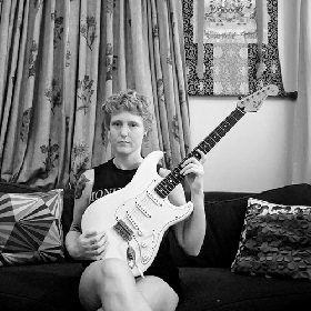 Person in black and white photo with tattoo on upper arm sits with legs crossed and a white electric guitar on their lap. They have short blond cropped hair and hold the neck of the guitar. In the background there is a decorative  full looking curtain with a floral pattern, they sit on a sofa with two patterned cushions