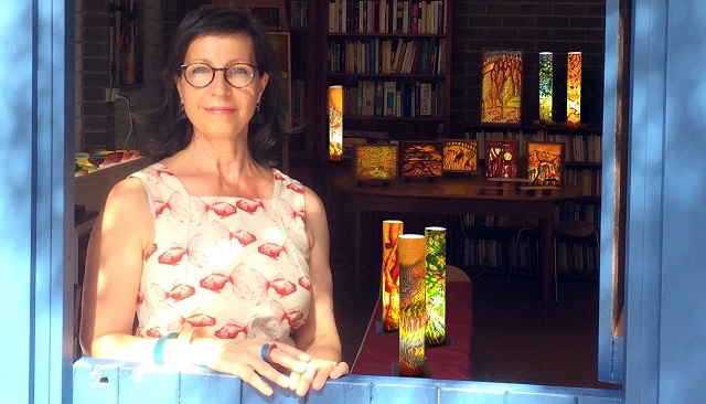 Photo of Rita inside her studio, ahe is framed by a stablke type light blue wooden door fram with her torso in view. In the background the image shows Rotas studio with books, light boxes, a table and chair. Rita wears a cream coloured singlet  with red fish on it, she as a blue and red ring, round glasses, a warm smile and dark shoulder length hair