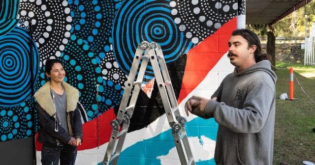 Photo of River Club Canoe Mural - photo shows a picture of the mural on the boat house wall, photo has image of two artists standing in front of the mural. Mural is bright green, blue, red and black. There are dots and blue like waves depicting the water and birdsy overlayed - picture is of incomplete art work.