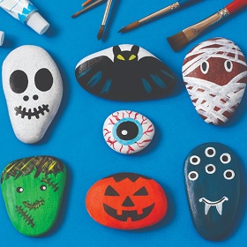 Blue background, placed on top are painted rocks, varying sizes and shapes with spooky motifs and iconography placed on the rocks. at the top of the image are 5 paintbrush tips and a tube of paint.