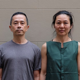Photo of artists Ryuichi Fujimura and WeiZen Ho stand side by side looking into the camera, they stand in front of a brown wall