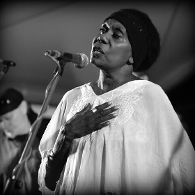 Black woman in white gown sings on stage wearing a black beanie, she has her hand on her chest and sings into a microphone.