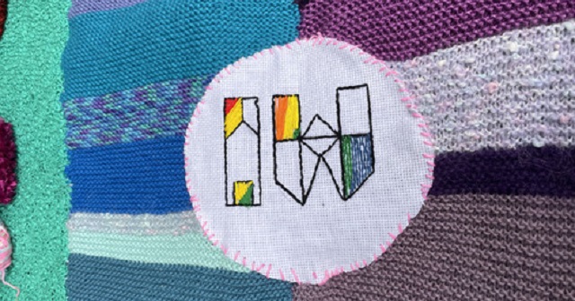 Photo of some hand made crochet, knitting and sewing combined together, the colours are purple, blue, green, white, pink, black, aqua, in the middle of the striped horizontal colour blocks is a sewn white circular patch attached with pink stitches, inside the patch is the inner west council logo with pride rainbow colours inlaid onto the logo "IW"