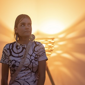Sammi Snedden Dunghutti & Wiradjuri wears woven earrings, she has a white tshirt with black flowers and swirls on the tshirt, behind her is out of focus with a sunset like hue, one of her woven baskets is in the background and casts a woven shadow against the wall