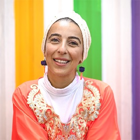 Photo of Sara Saleh, Sara smiles at the camera she has a white turban on and black cube drop earrings, she wears a orange and gold kaftan and a white turtle neck underneath
