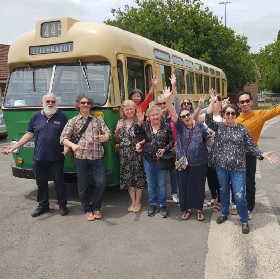 Group of people photographed in front of a vintage green and yellow bus, they wave at the camera