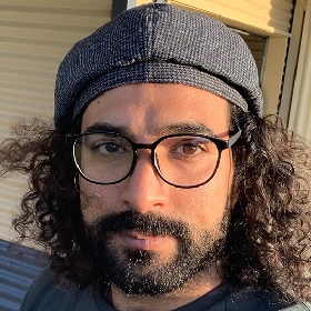 Photo of a person with black tshotr, beard and curly shoulder length hair they wear a grey textured beret and black framed round glasses