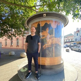 Simon Greiner stands infront of the public art installation Newtown Art Seat, his work is captured inside the glass cylindrical display and features a photograph of a cardboard dog that looks like it is part of a merry go round