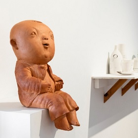 Clay sculpture of a chubby rounded face child like figure sitting on the side of a plinth, the clay is terracotta colour
