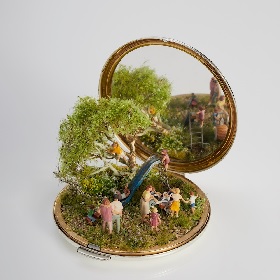 Photo of an open gold rimmed pocket mirror inside is a miniature sculptural artwork featuring a park scene with a large tree, a slide and a group of children and adults